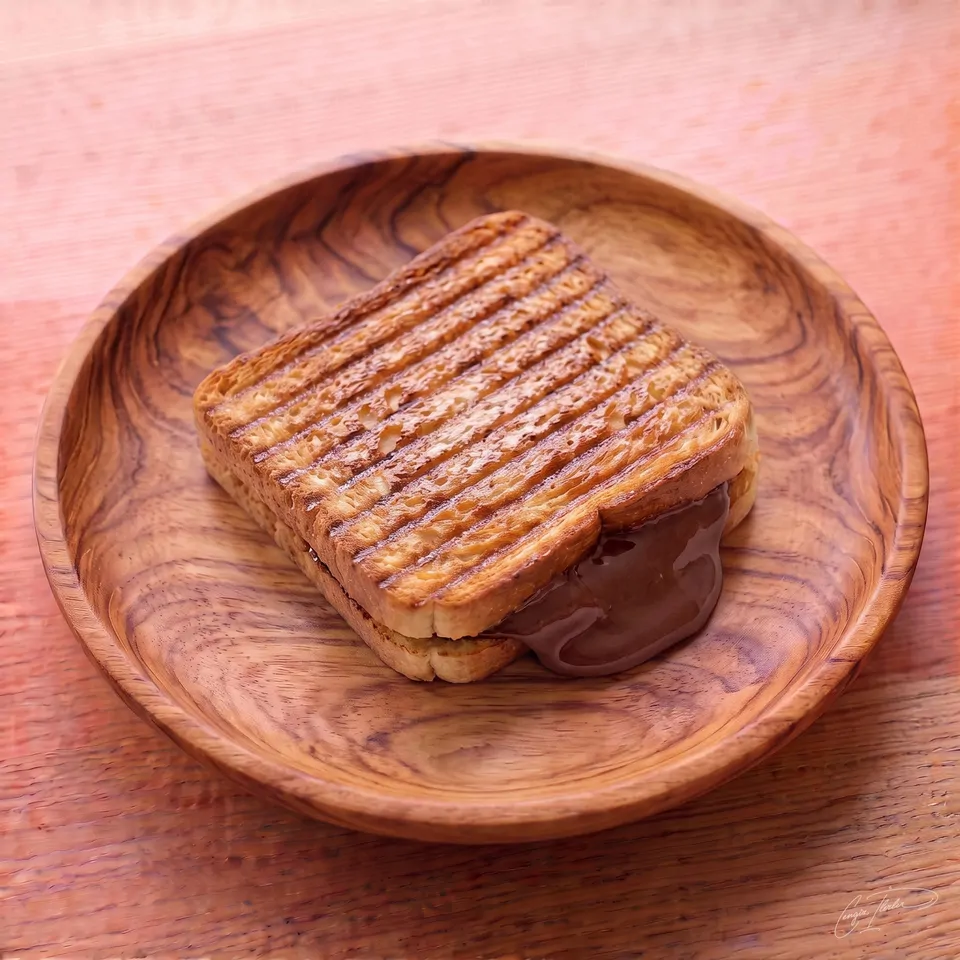 A perfectly grilled çikolatalı tost (chocolate toast) on a carved wooden plate, with deep horizontal grill marks on the golden-brown bread and warm, melted chocolate hazelnut spread oozing out the side.