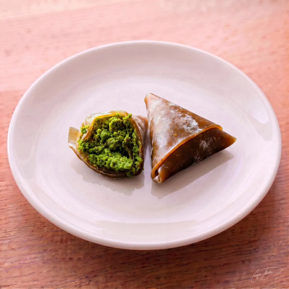 Two triangle-shaped muska (amulet) treats on a white plate, featuring a translucent, dark fruit-leather (pestil) wrapper folded over a dense, bright green pistachio filling.