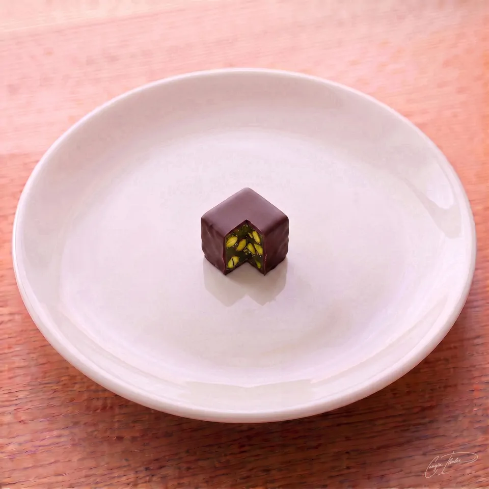 A single cube of chocolate-covered lokum on a white plate, with a corner cut away to reveal a vibrant green, pistachio-filled Turkish delight interior.