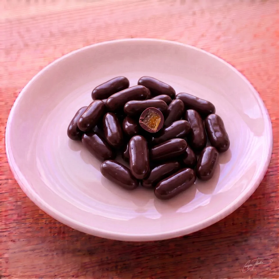 A small pile of dark chocolate-coated orange dragees on a white plate, with one piece cut open to reveal a bright orange candied center.