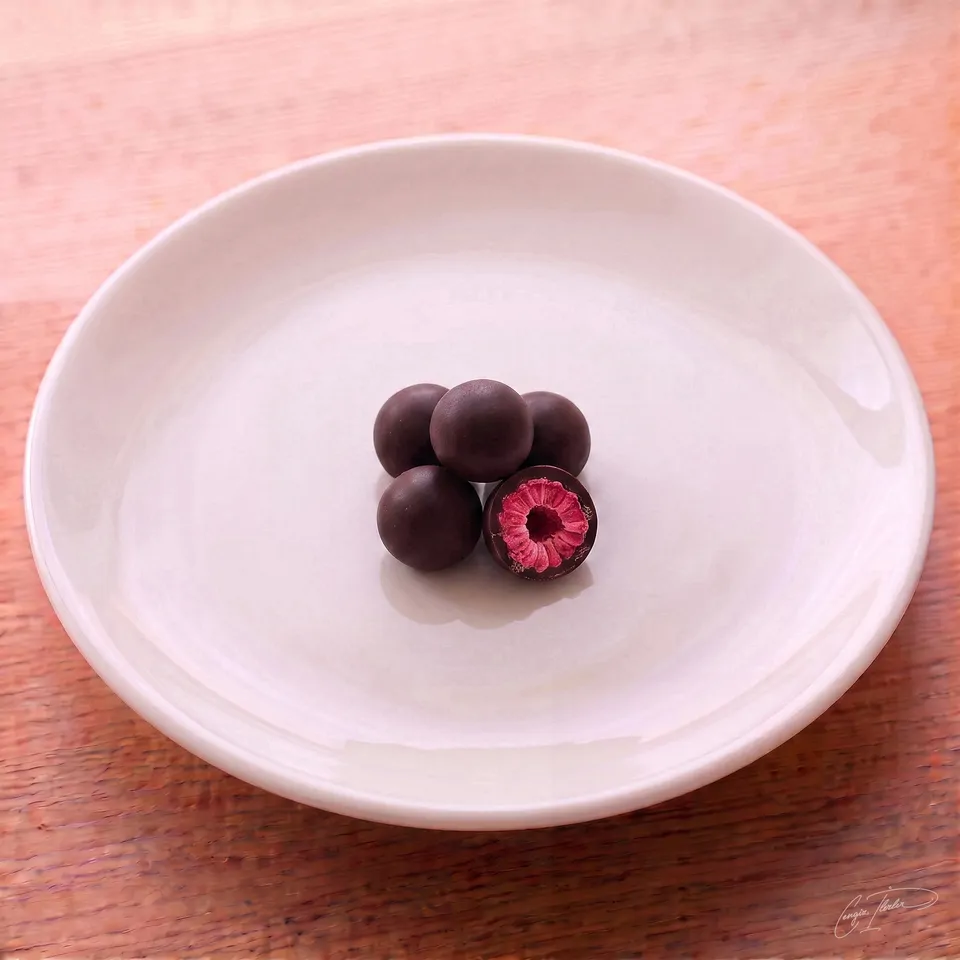 A small pile of dark chocolate-coated raspberry dragees on a white plate, with one piece cut open to reveal a bright pink, textured raspberry center.