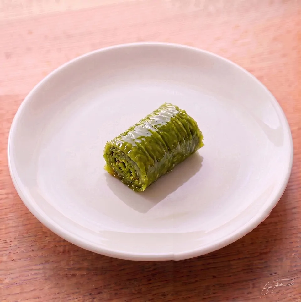 A single piece of fıstıklı dolama — a vivid green, tightly rolled Turkish dessert made almost entirely of ground pistachios wrapped in a single layer of phyllo dough and soaked in syrup — on a white plate.