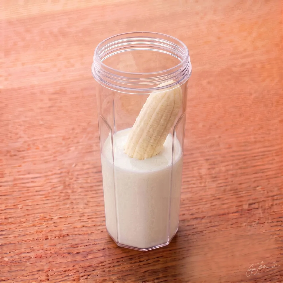 A clear blender cup containing fresh milk and a half-peeled banana standing upright inside it, resting on a wooden table.