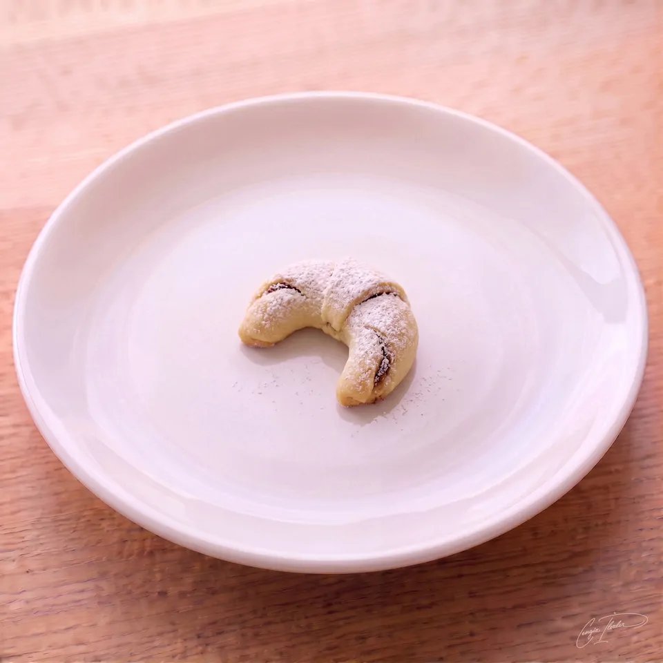 A single crescent-shaped elmalı kurabiye — a soft Turkish apple cookie dusted generously with powdered sugar — resting on a white plate.