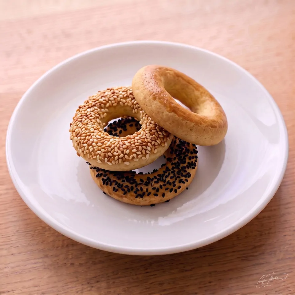 A stack of three small kandil simidi rings on a white plate — one plain, one coated in sesame seeds, and one coated in black nigella seeds.