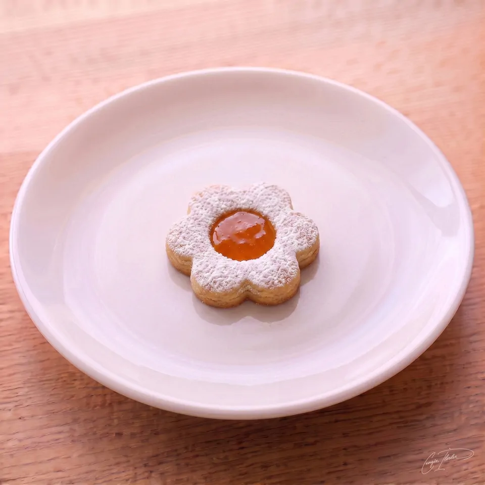 A single flower-shaped shortbread cookie dusted with powdered sugar, with a bright orange marmalade center, resting on a white plate.