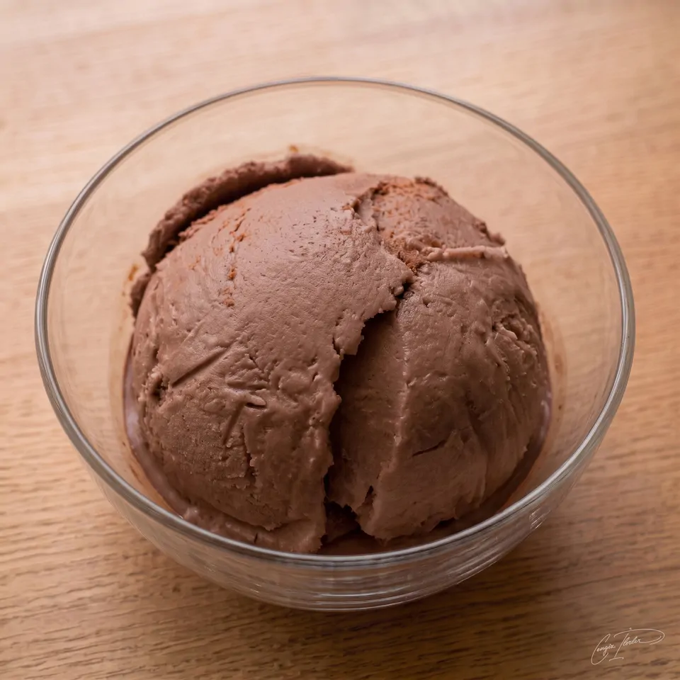 A single scoop of glossy chocolate ice cream in a clear glass bowl on a wooden table.