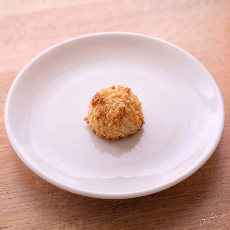 A single golden-brown coconut macaroon — a domed mound of baked shredded coconut — on a white plate on a wooden table.