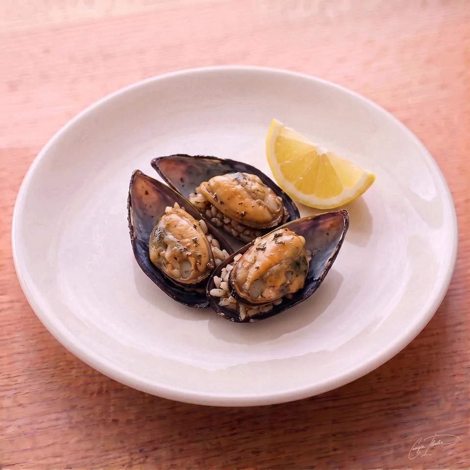 A white plate holding three midye dolma (stuffed mussels) — their dark shells opened to reveal a savory, spiced rice filling — served with a wedge of fresh lemon.