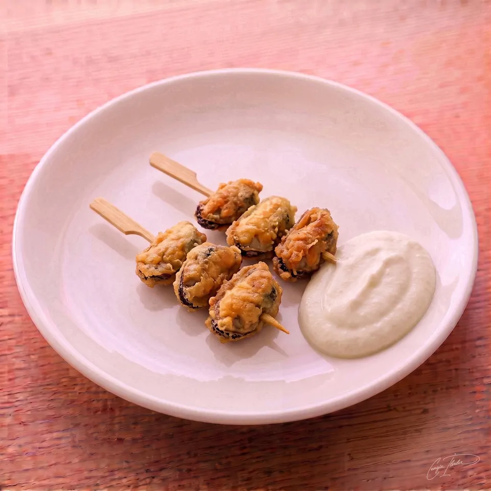 Two wooden skewers of golden, crispy midye tava (fried mussels) resting on a white plate beside a generous dollop of creamy tarator sauce.